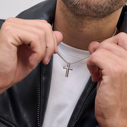 Man wearing a silver cross necklace, holding the pendant in his hands, dressed in a white shirt and black leather jacket.